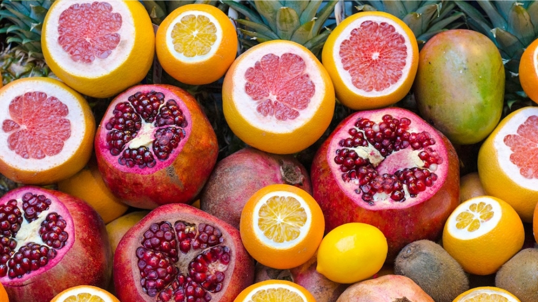 A vibrant assortment of cut and whole citrus fruits and pomegranates, showcasing a range of oranges, yellows and deep reds, with a backdrop of green foliage.