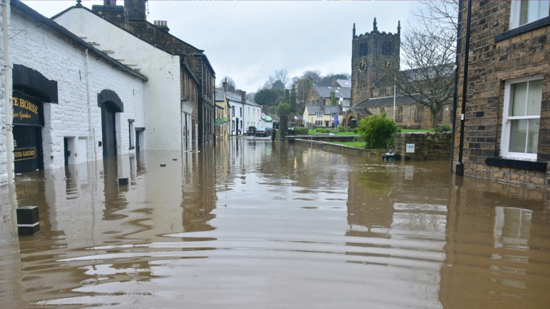 A flooded street in a village with water levels reaching the doors of traditional stone buildings, and a church tower visible in the background under an overcast sky.