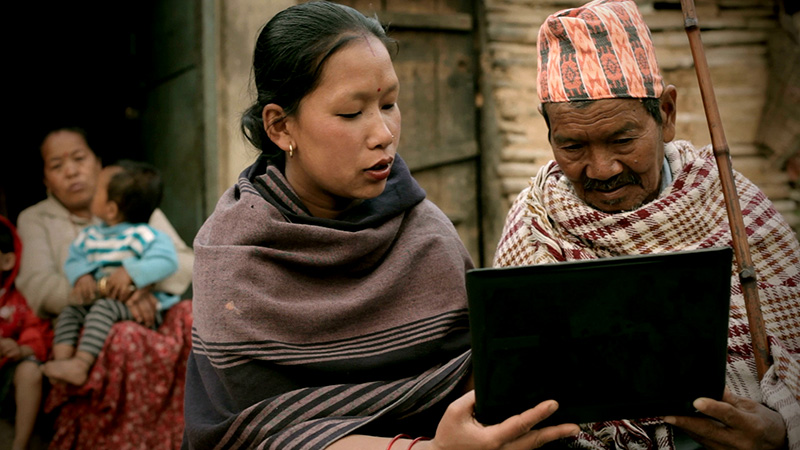 A woman and an older man engage with a laptop, with the woman holding the device and explaining, while the man looks on attentively. In the background, a woman sits cradling a child.