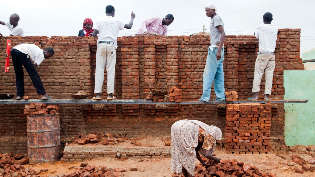 Several people are working on constructing a brick wall. The wall is partially built, with stacks of red bricks visible.