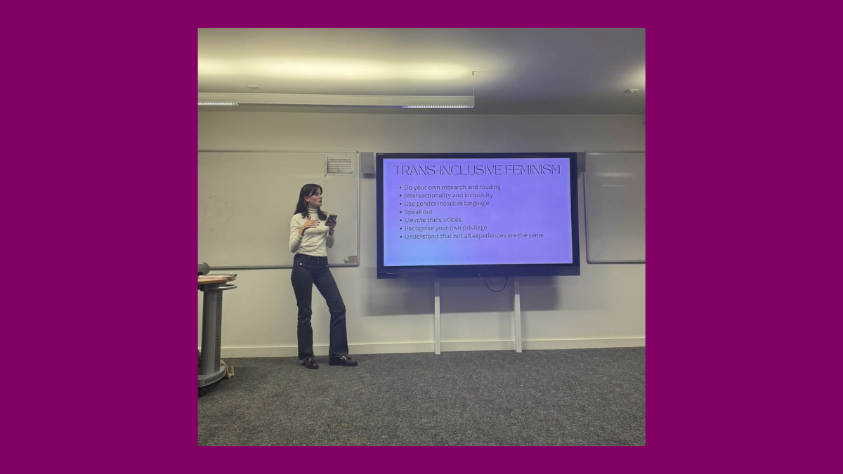 Photo of Ione Gildroy standing next to a screen that reads 'Trans Inclusive Feminism' and a list of bullet points on how to act in accordance with trans inclusive feminism.