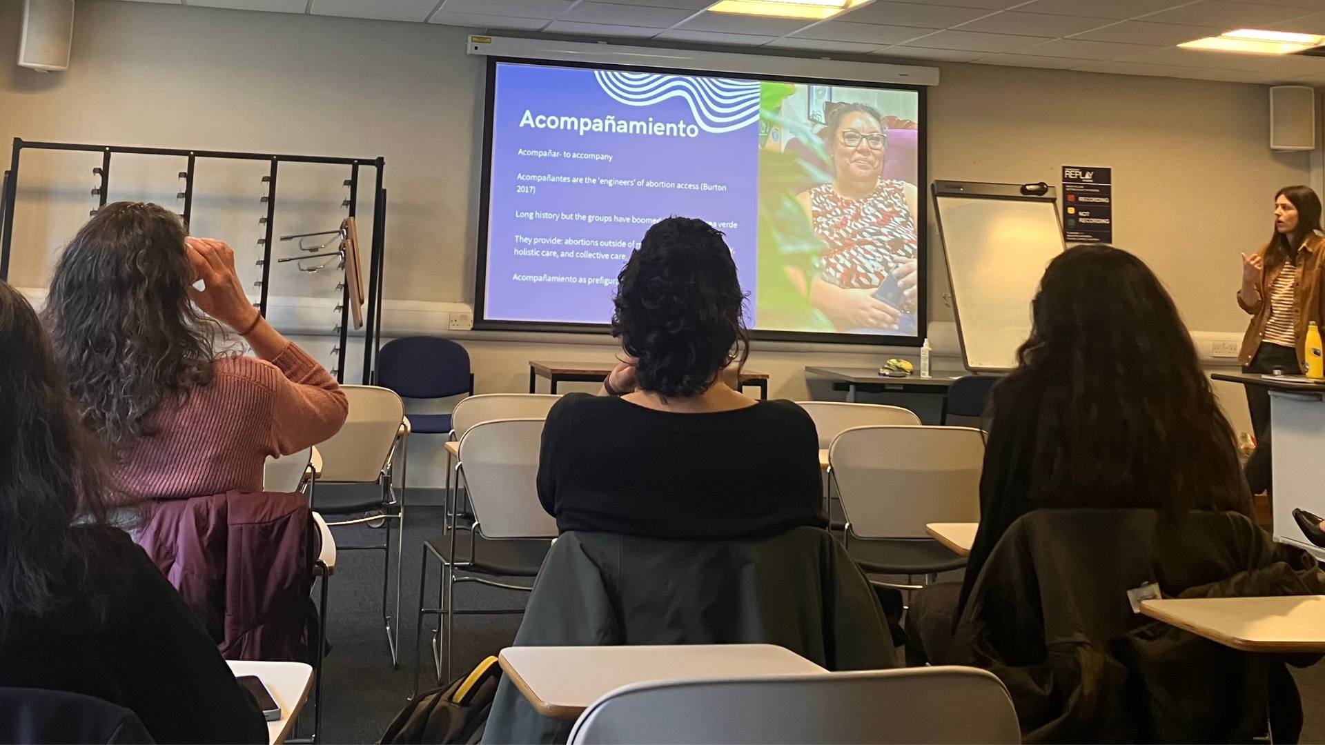 Photo of Cordelia Freeman at the event speaking in front of a group of people. Next to her is a screen that reads 'Accompañamientos' and on the right side of the screen, there is a photo of an activist, who is smiling and wearing glasses and a top with an abstract pattern.