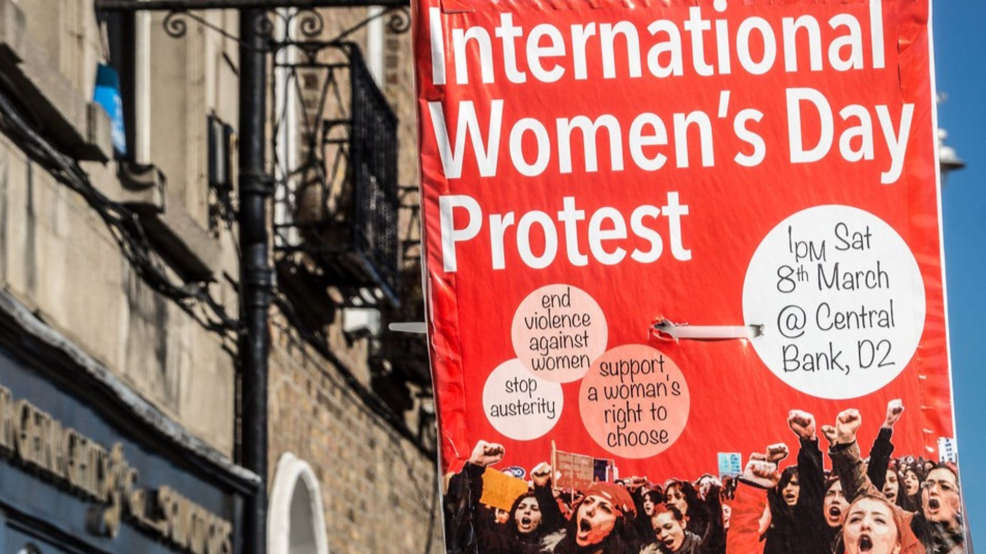 Photo of a poster on a lamp post in a street that reads 'International Women's Day Protest'. There is an image of people protesting and chanting under the text on the poster.