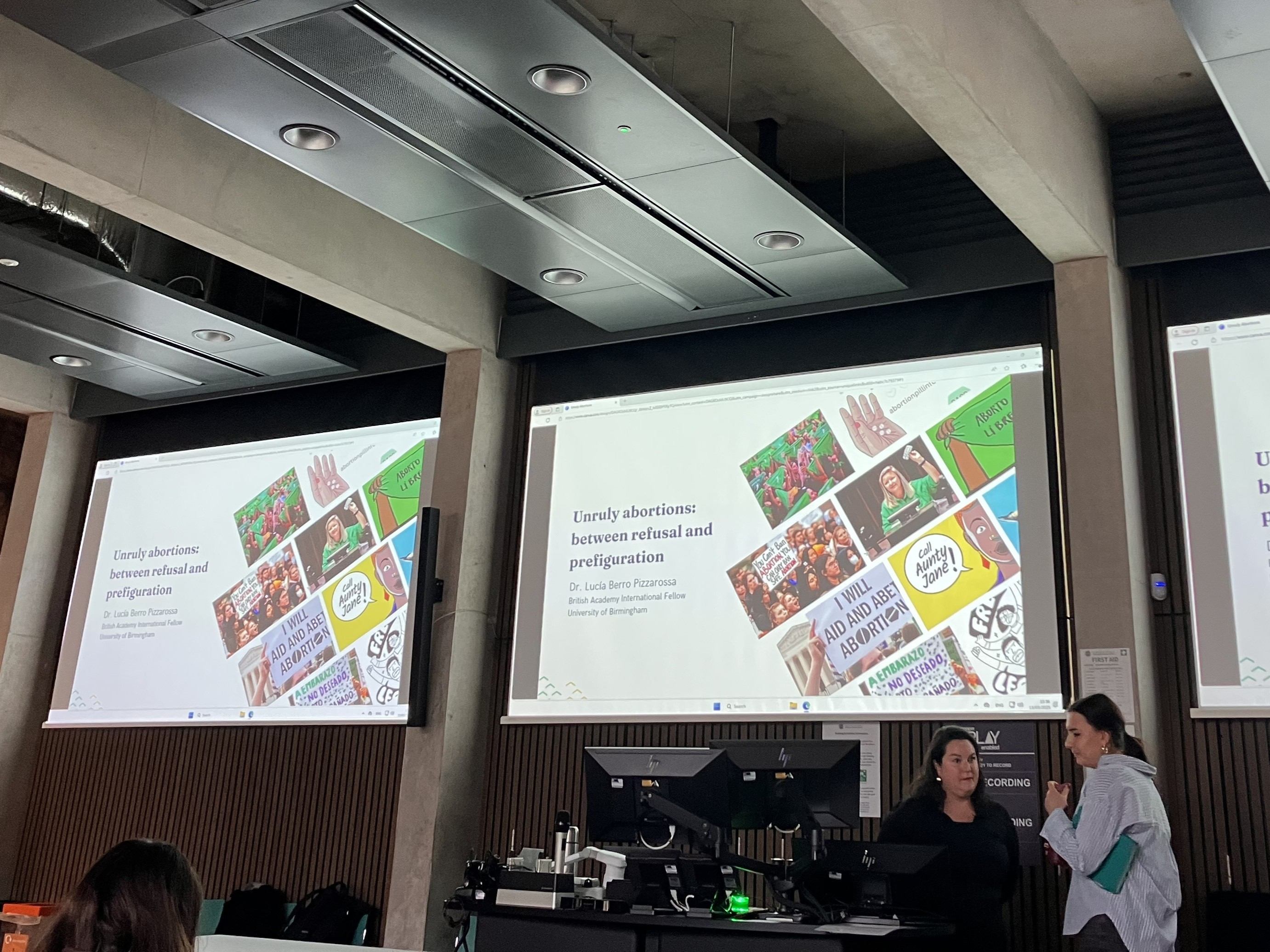 Photo of Dr Lucía Berro Pizzarossa and another person talking next to two screens before the lecture. Behind them are three screens that read 'Unruly abortions: Between refusal and prefiguration'. The screen also has photos of slogans/posters by self-managed abortion collectives.