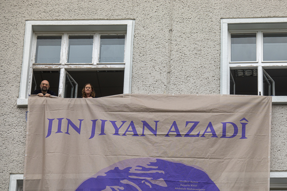 Two people holding a banner out of a window, with a women on the banner.