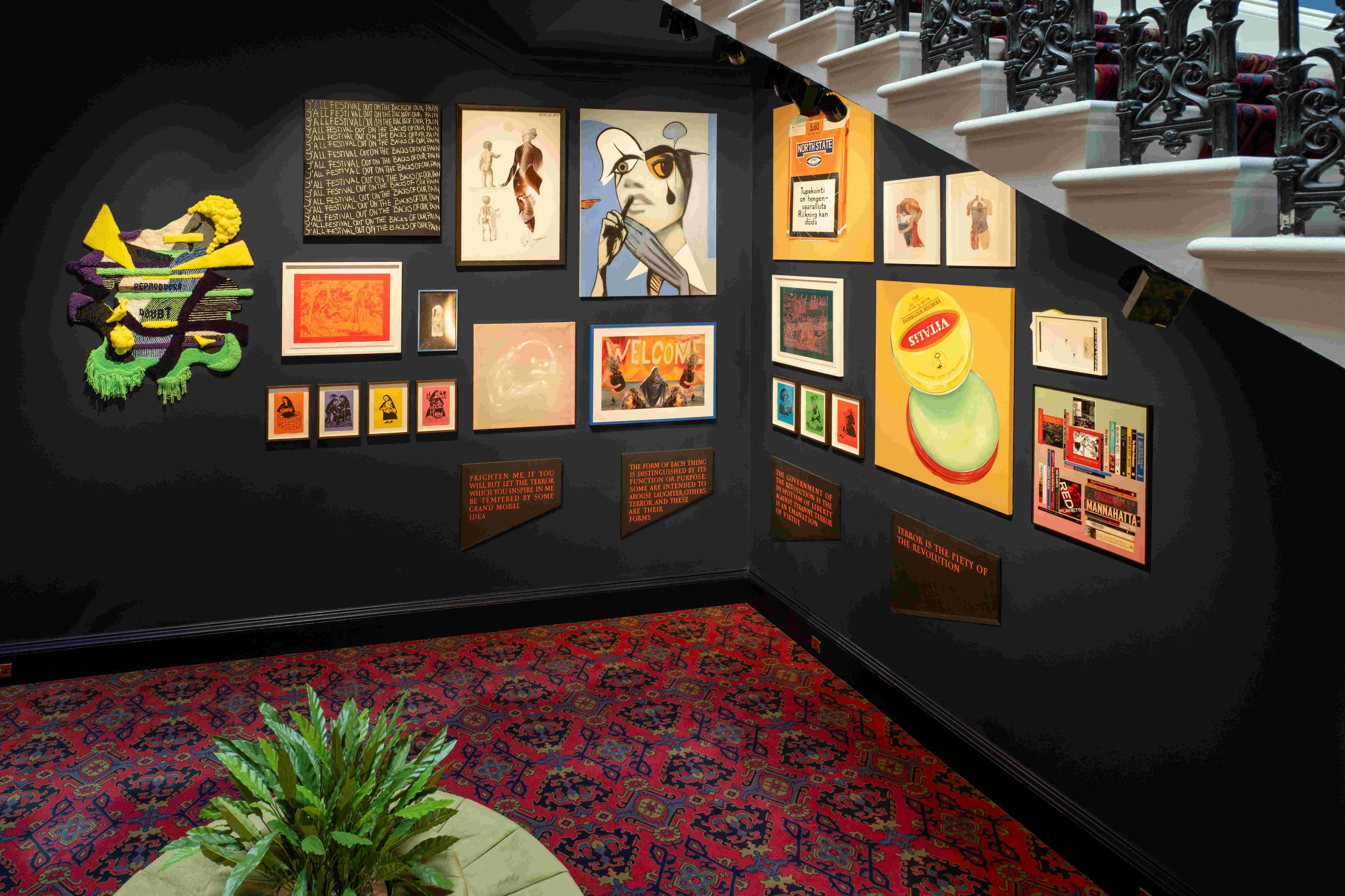 Display of art works at the Old College of the University of Edinburgh. The display is located below a staircase and consists of a variety of vibrant and colourful pieces. There is a plant across the artwork. 