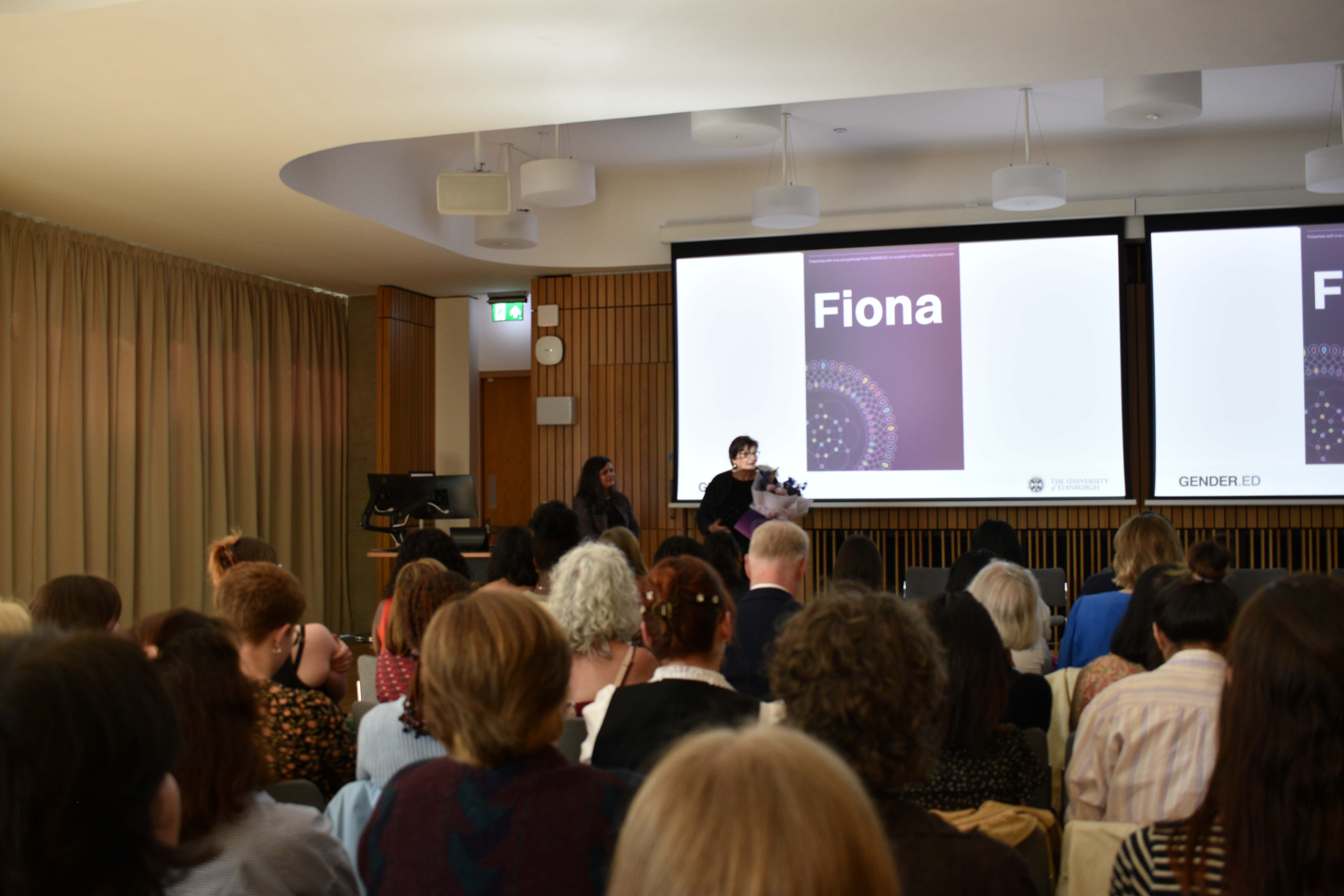 Photo of Radhika Govinda and Fiona Mackay in front of rows of people at the Annual Research Showcase. Fiona is holding a bouquet of flowers in her hand and behind her is a screen that reads 'Fiona' and a mandala.