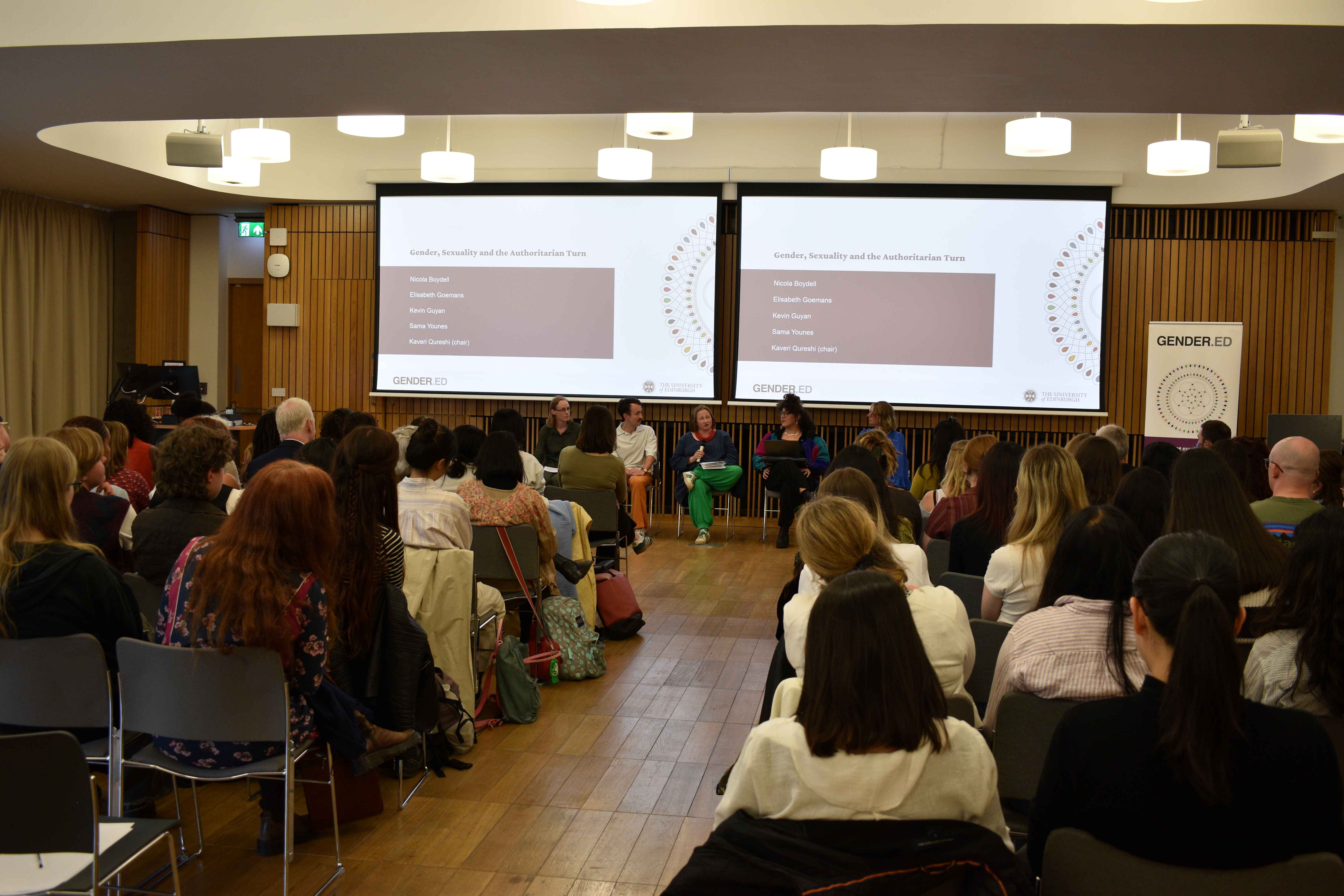 A photo of the panelists at the panel discussion. They are seated in a row in front of an audience. Behind them are two screens with slides that have their names listed.