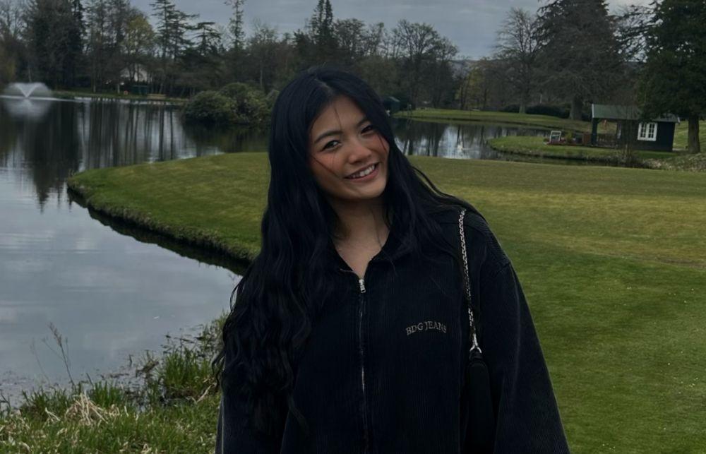 A picture of Maggie next to a lake with trees in the background. Maggie wears a black zip-up hoodie