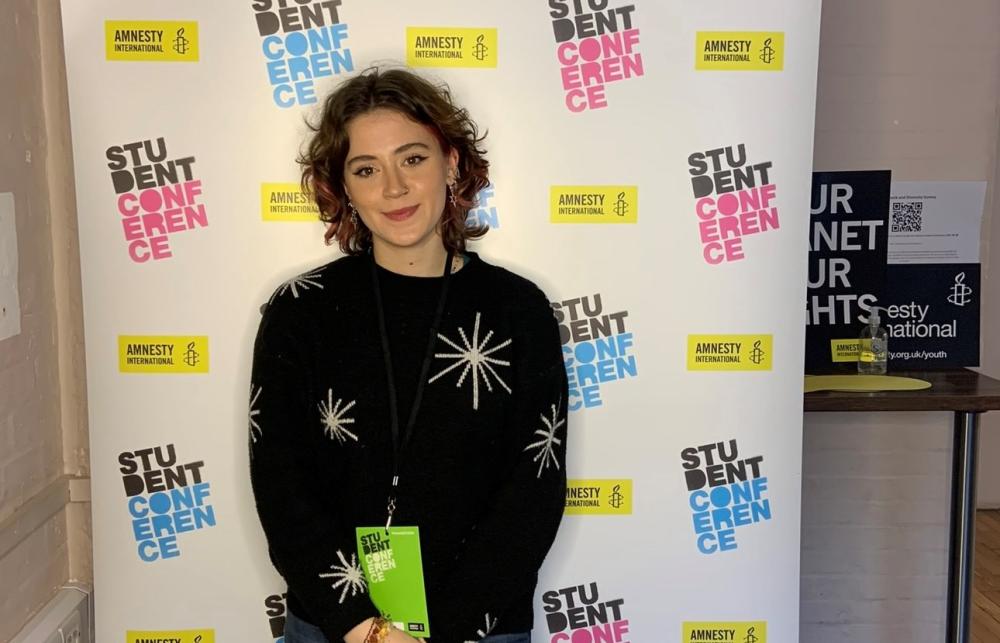 Picture of Kate Wilson. They are smiling, wearing a jumper with stars on it. Behind them is a roller banner that reads 'Amnesty International' and 'Student Conference'