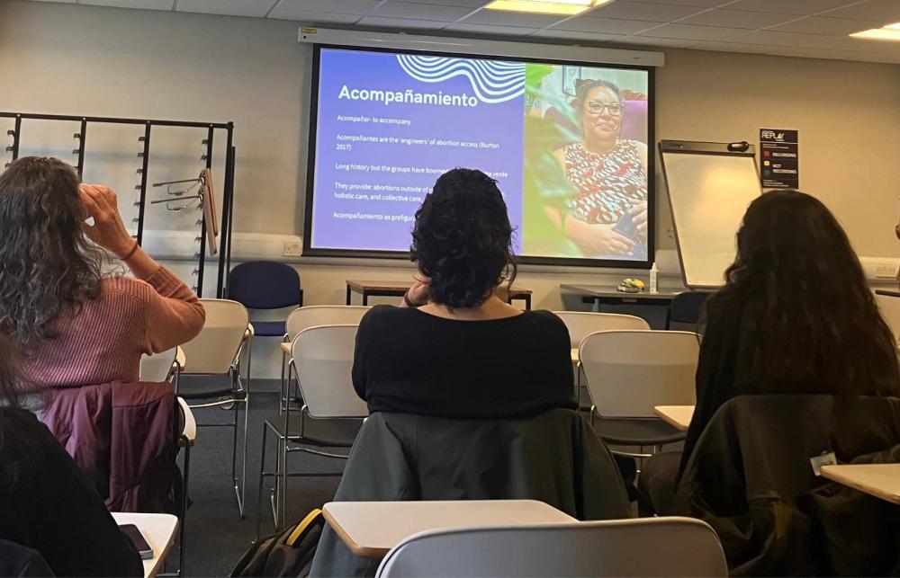 Photo of Cordelia Freeman at the event speaking in front of a group of people. Next to her is a screen that reads 'Accompañamientos' and on the right side of the screen, there is a photo of an activist, who is smiling and wearing glasses and a top with an abstract pattern. 