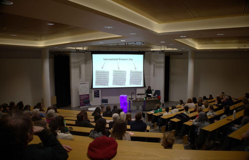 Photo of Naeema Yaqoob Sajid giving the International Women's Day Lecture 2025 in a lecture hall with an audience. Behind her is a screen that reads 'International Women's Day'. People are 