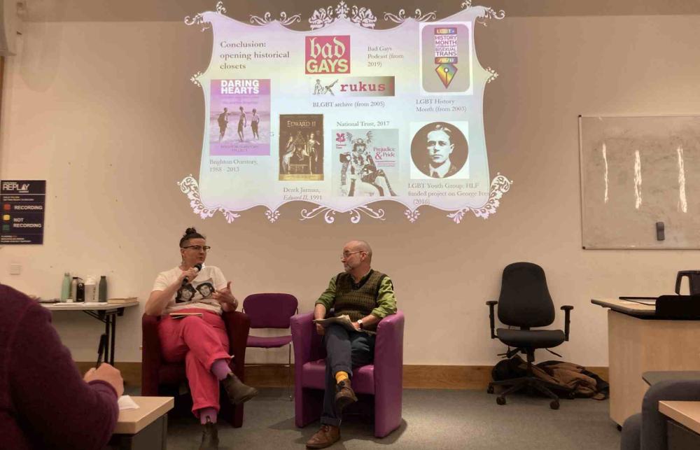 Photo of Professor Matt Cook and the panelists in Meadows Lecture Theatre sitting on two chairs, having a conversation. Behind them there is a screen with various images of podcasts, archives, and projects on 'opening the historical closet'.