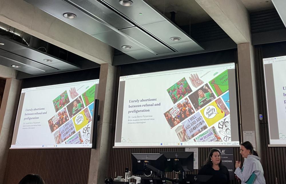 Photo of Dr Lucía Berro Pizzarossa and another person talking next to two screens before the lecture. Behind them are three screens that read 'Unruly abortions: Between refusal and prefiguration'. The screen also has photos of slogans/posters by self-managed abortion collectives. 
