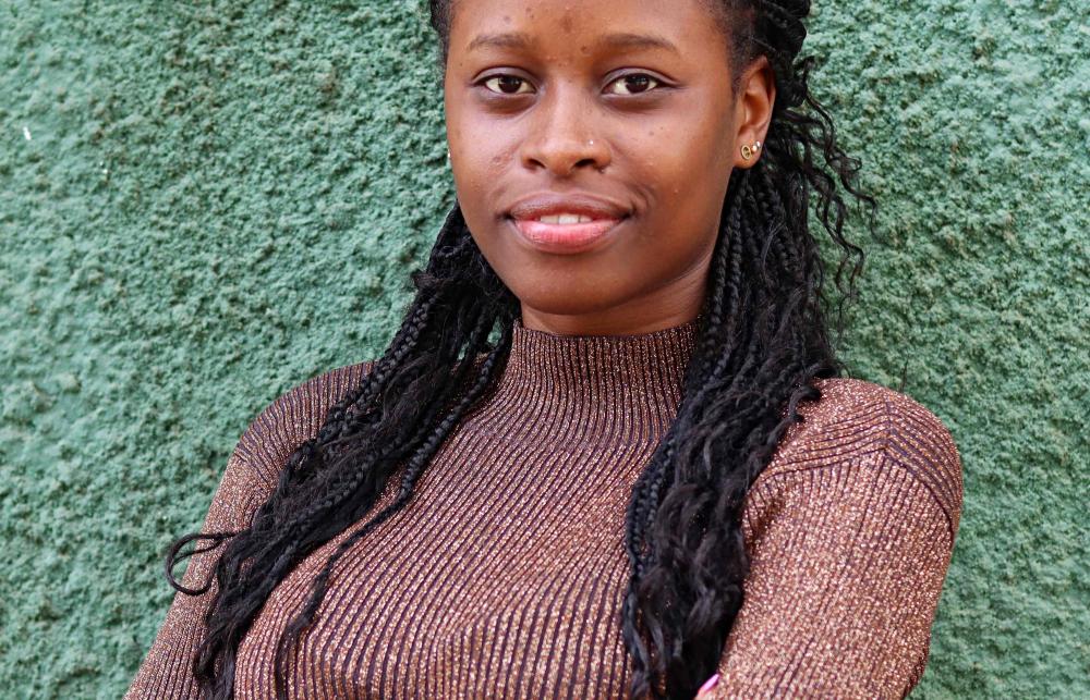 Portrait of Maria Tsuma smiling. She is wearing a sparkly turtleneck jumper and has her arm crossed.