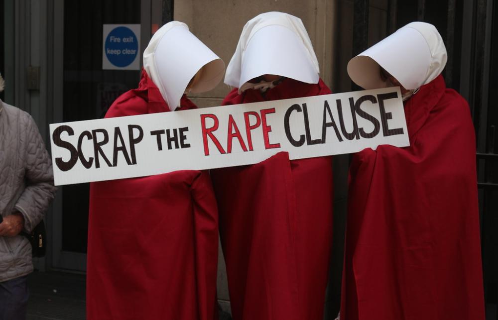 Three people dressed as 'Handmaids' from Margaret Atwood's 'The Handmaid's Tale'. They are wearing red long robes and white head wear that covers their hair and faces. They are holding a piece of paper that reads 'Scrap the Rape Clause'