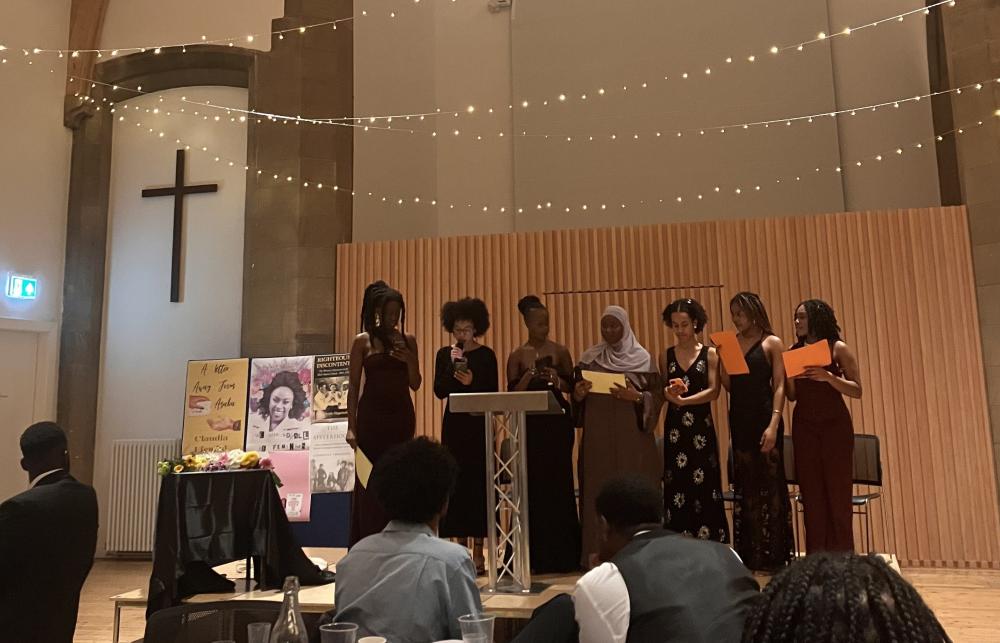 7 members of Black Women* at Edinburgh's Committee on a stage at the Magazine Launch Event. Above them are some strings of fairy lights. Behind and to the left of them, there is a cross and some posters. One of the committee members, Maryam Yusuf, is speaking into a microphone. There is an audience across the stage.