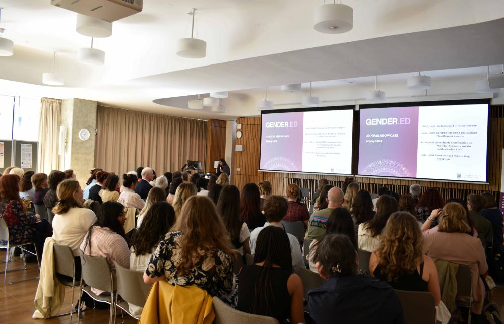 Photo of Radhika Govinda, director of GENDER.ED, speaking in front of rows of people at the Annual Research Showcase. She is holding a microphone. To her right, there is a screen that with a slide that reads 'GENDER.ED'.