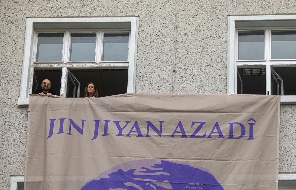 Two people holding a banner out of a window, with a women on the banner.