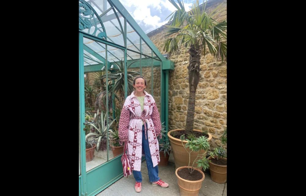 Photo of Emily Clifford in a long pink and white coat next to a large plant and greenhouse
