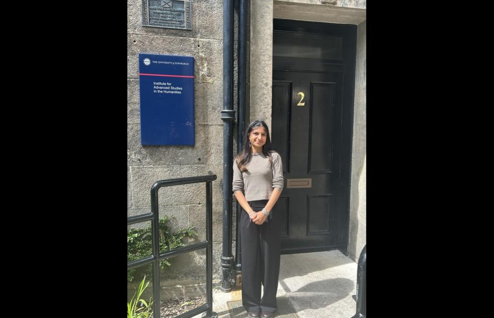Photo of Jayni Makwana standing with her hands clasped in front of the main door at the IASH building on the university campus
